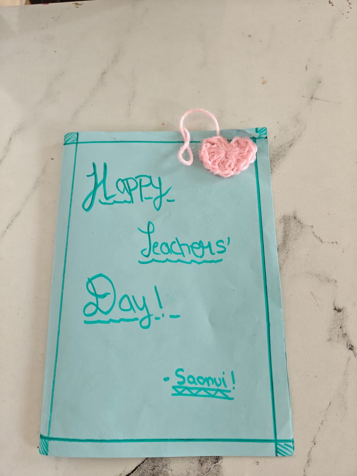 Saanvi's Teacher's Day card: sea-green paper with a ruled border, Happy Teachers' Day written in marker, and a small pink crochet heart tied on with yarn
