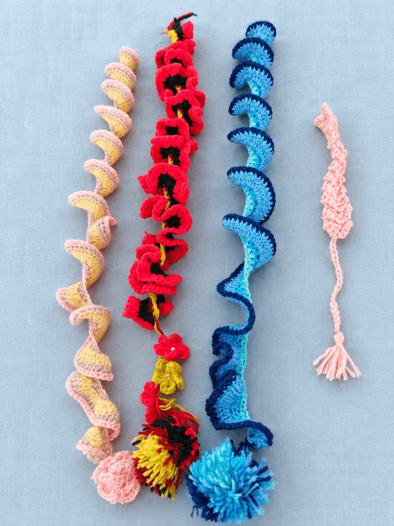 Four long crochet spirals: one in pink and yellow with a pink ball at the end, one covered in red and black ruffled flowers with a yellow tassel, one in shades of blue with a blue pompom, and a slim pink braided piece with a tassel